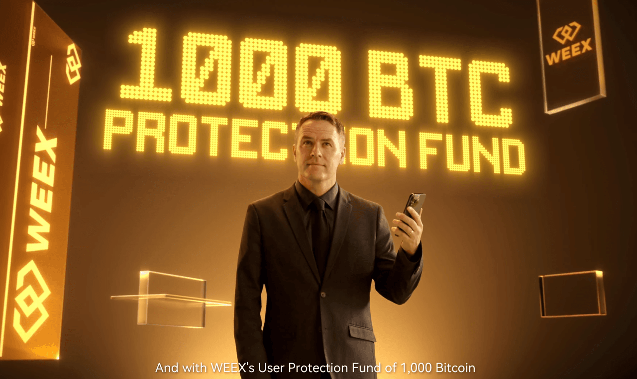 Michael Owen đứng trong một căn phòng vàng với điện thoại thông minh trên tay. Phía sau anh ấy, dòng chữ “1000 BTC Protection Fund” phát sáng bằng chữ vàng lớn, được bao quanh bởi các logo WEEX.